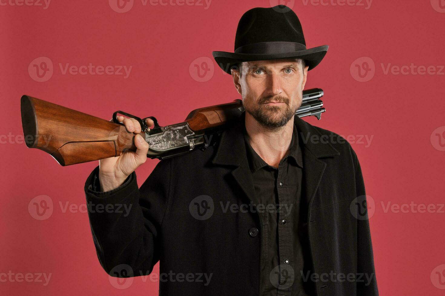 Middle-aged man with beard, mustache, in black jacket and hat, holding a gun while posing against a red background. Sincere emotions concept. photo