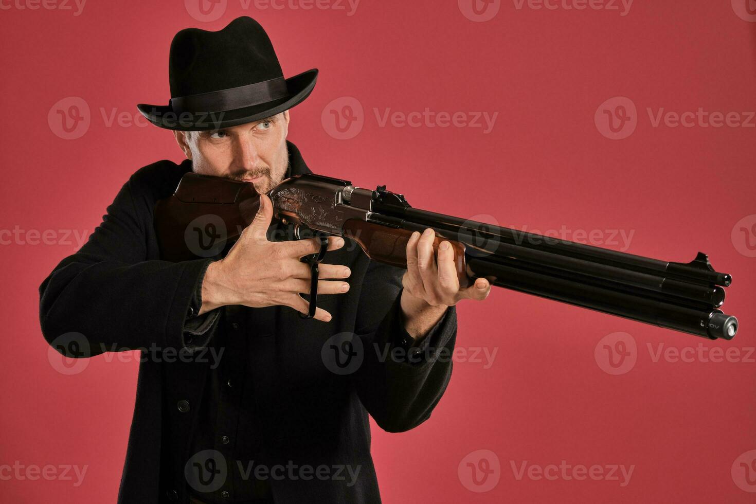 Middle-aged man with beard, mustache, in black jacket and hat, holding a gun while posing against a red background. Sincere emotions concept. photo