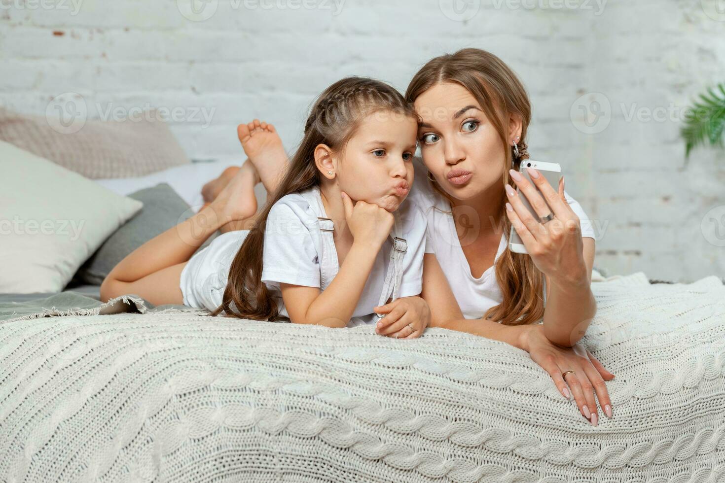 Indoor portrait of a beautiful mother with her charming little daughter posing against bedroom interior. photo