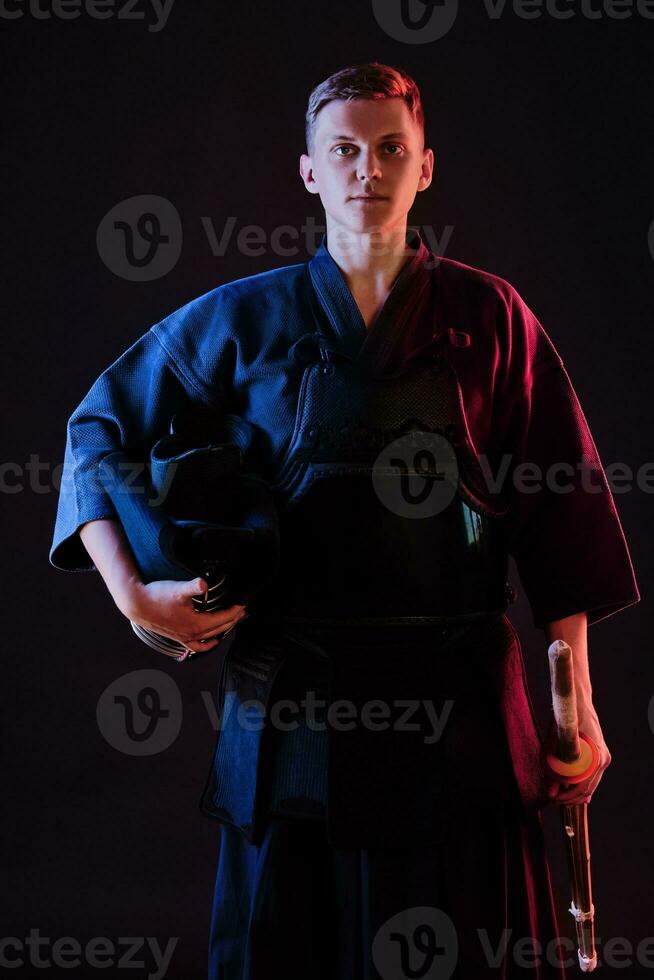 Kendo fighter wearing in an armor, traditional kimono is holding his helmet and shinai bamboo sword while posing on a black background. Close up. photo