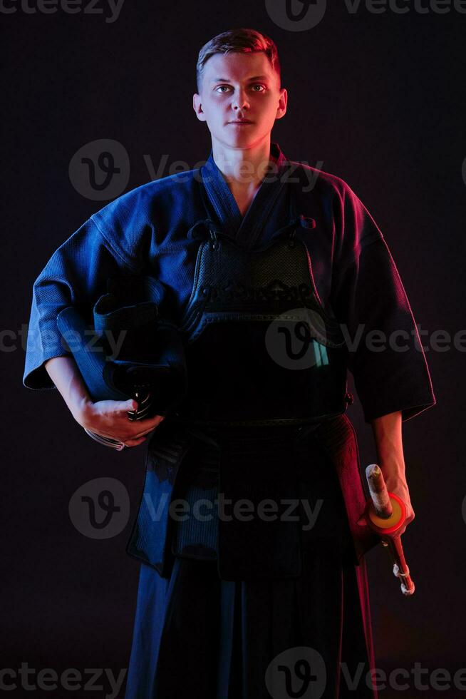 Kendo fighter wearing in an armor, traditional kimono is holding his helmet and shinai bamboo sword while posing on a black background. Close up. photo