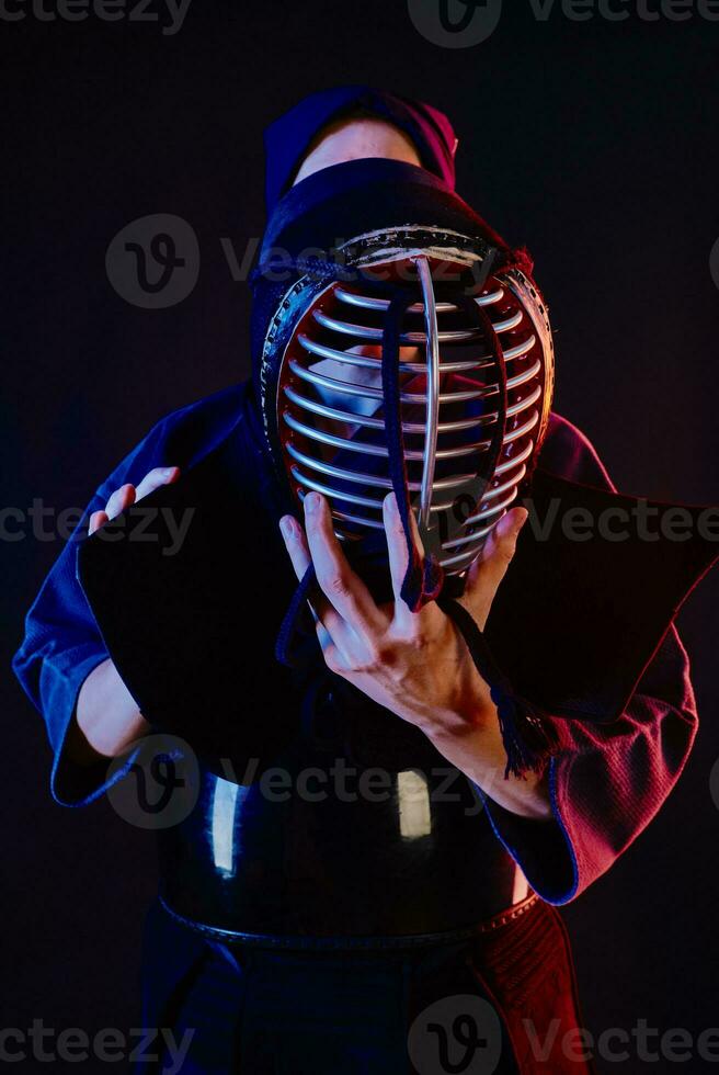 cerca arriba disparo, kendo combatiente vistiendo en un armadura y tradicional kimono es atadura el cordones en su casco en pie en contra un negro antecedentes. foto