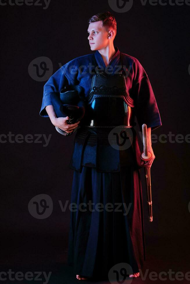 Kendo fighter wearing in an armor, traditional kimono is holding his helmet and shinai bamboo sword while posing on a black background. Close up. photo