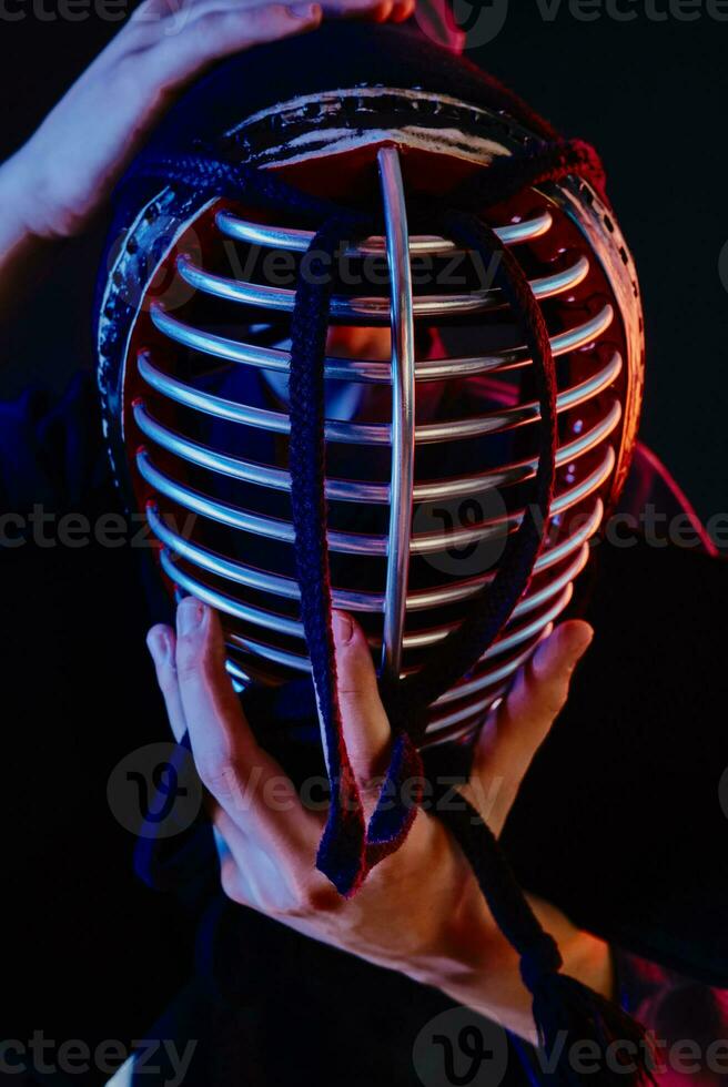 Close up shot, Kendo fighter wearing in an armor and traditional kimono is tying the lacing on his helmet standing against a black background. photo
