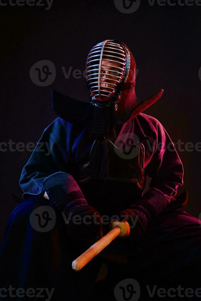 Close up. Kendo fighter wearing in an armor, traditional kimono, helmet, sitting, practicing martial art with shinai bamboo sword, black background. photo