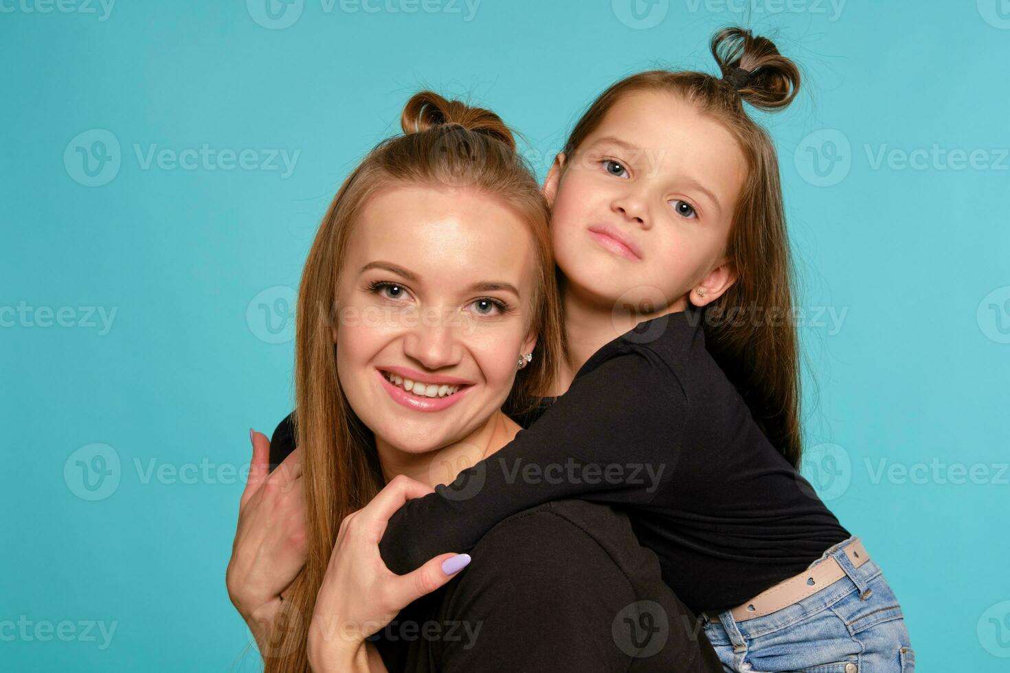 Mom and daughter with a funny hairstyles, dressed in black shirts and blue denim jeans are posing against a blue studio background. Close-up shot. photo