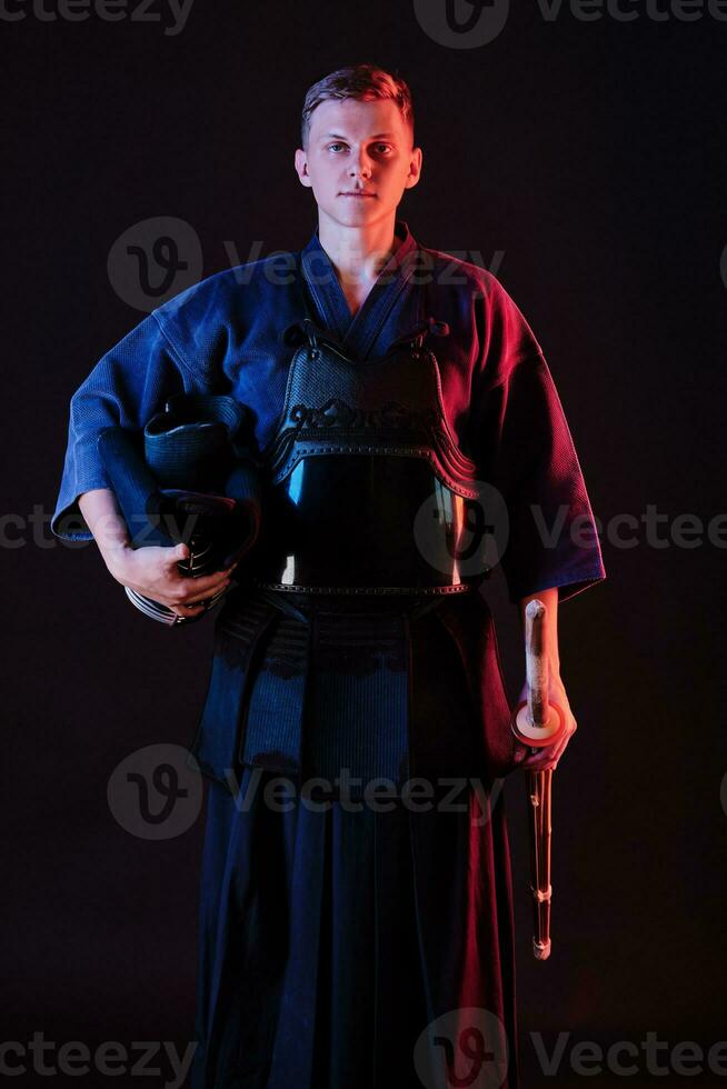 Kendo fighter wearing in an armor, traditional kimono is holding his helmet and shinai bamboo sword while posing on a black background. Close up. photo