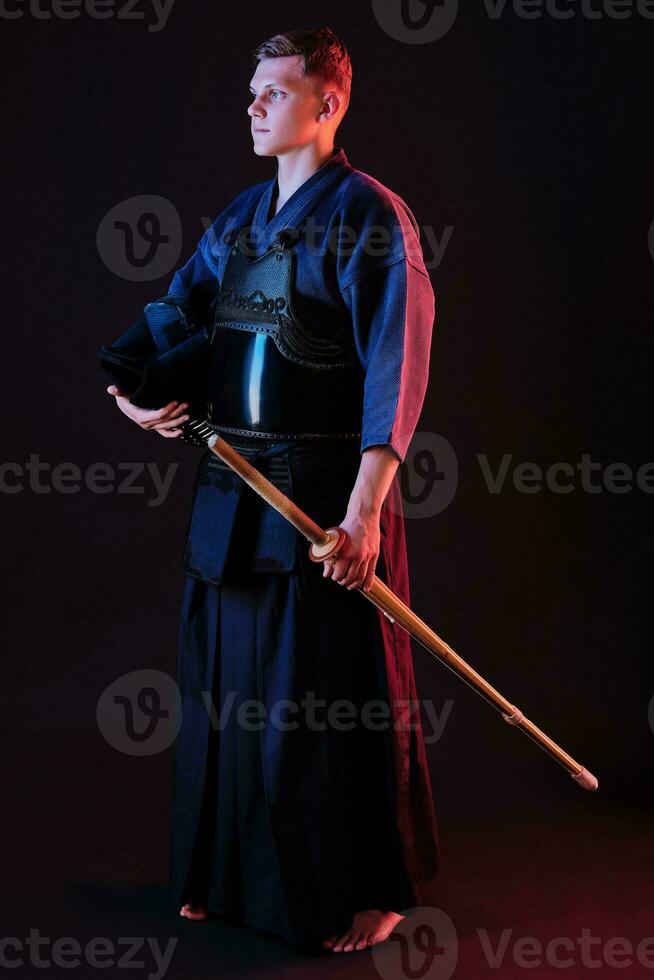 Kendo fighter wearing in an armor, traditional kimono is holding his helmet and shinai bamboo sword while posing on a black background. Close up. photo