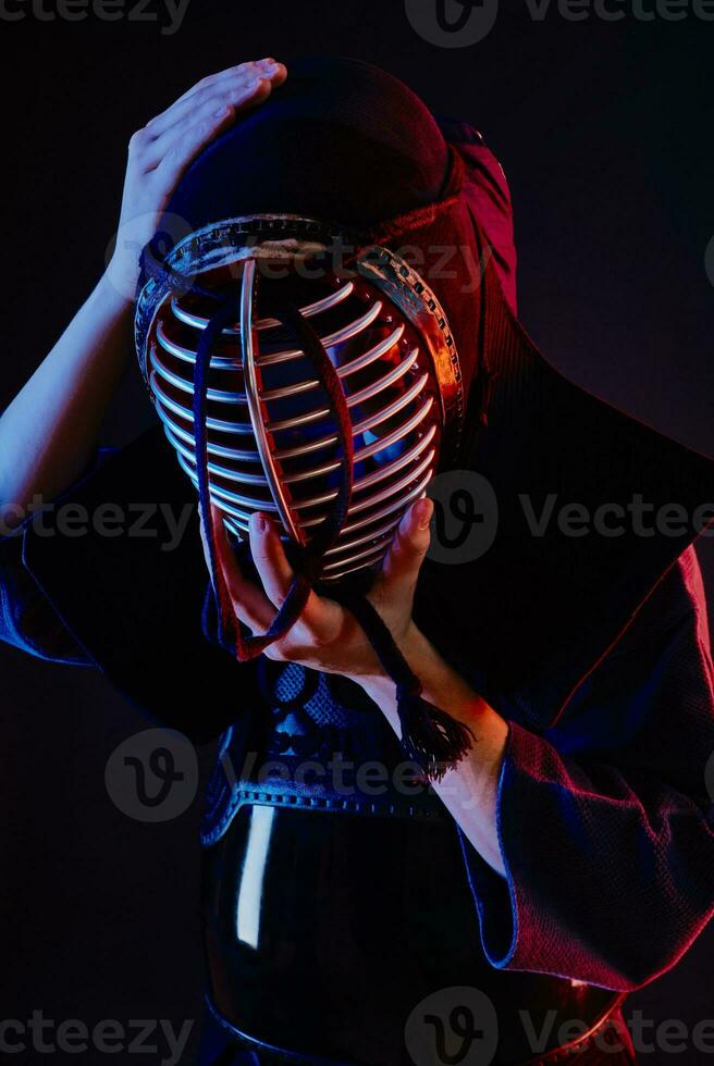 cerca arriba disparo, kendo combatiente vistiendo en un armadura y tradicional kimono es atadura el cordones en su casco en pie en contra un negro antecedentes. foto