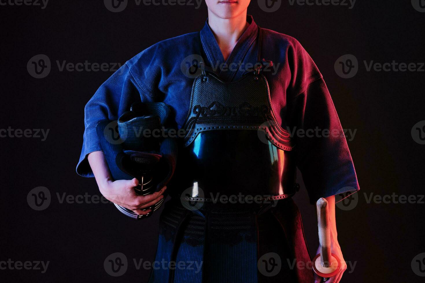 Kendo fighter wearing in an armor, traditional kimono is holding his helmet and shinai bamboo sword while posing on a black background. Close up. photo