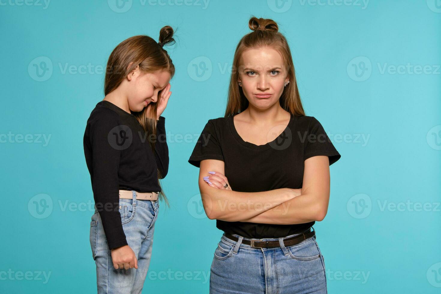 Mom and daughter with a funny hairstyles, dressed in black shirts and blue denim jeans are posing against a blue studio background. Close-up shot. photo