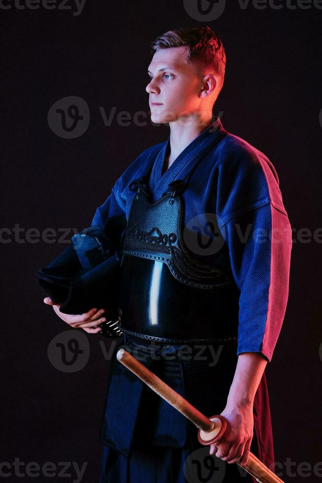 Kendo fighter wearing in an armor, traditional kimono is holding his helmet and shinai bamboo sword while posing on a black background. Close up. photo