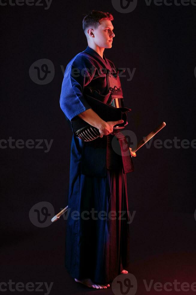 Kendo fighter wearing in an armor, traditional kimono is holding his helmet and shinai bamboo sword while posing on a black background. Close up. photo