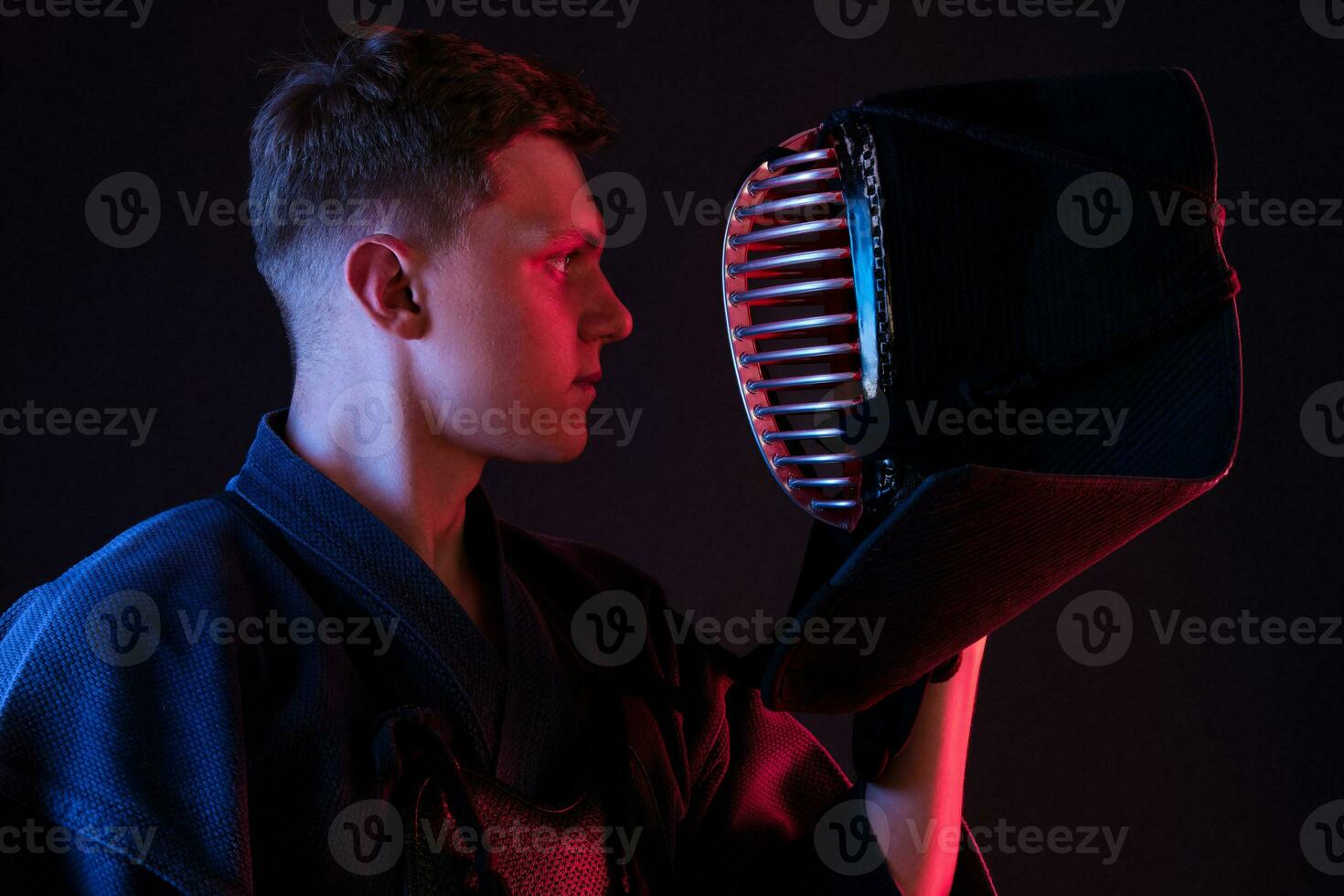 Close up shot, Kendo fighter wearing in an armor, traditional kimono is looking at his helmet while standing against a black studio background. photo