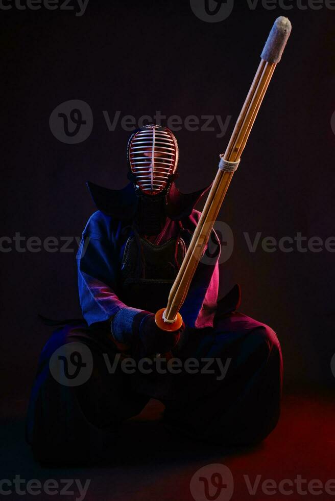 Close up. Kendo fighter wearing in an armor, traditional kimono, helmet, sitting, practicing martial art with shinai bamboo sword, black background. photo