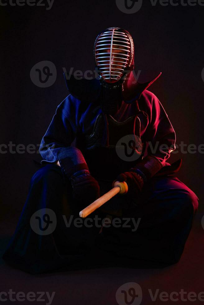 Close up. Kendo fighter wearing in an armor, traditional kimono, helmet, sitting, practicing martial art with shinai bamboo sword, black background. photo