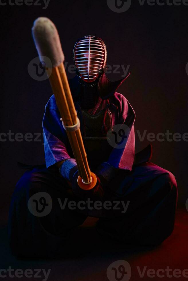 Close up. Kendo fighter wearing in an armor, traditional kimono, helmet, sitting, practicing martial art with shinai bamboo sword, black background. photo