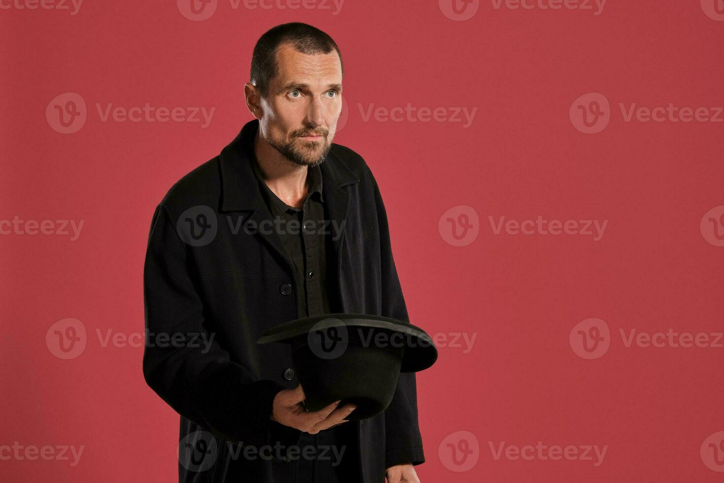 de edad mediana hombre con barba y Bigote, usa negro sombrero y chaqueta posando en contra un rojo antecedentes. sincero emociones concepto. foto