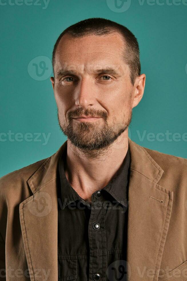 Middle-aged man with beard and mustache, wears black shirt and brown jacket posing against a blue background. Sincere emotions concept. photo
