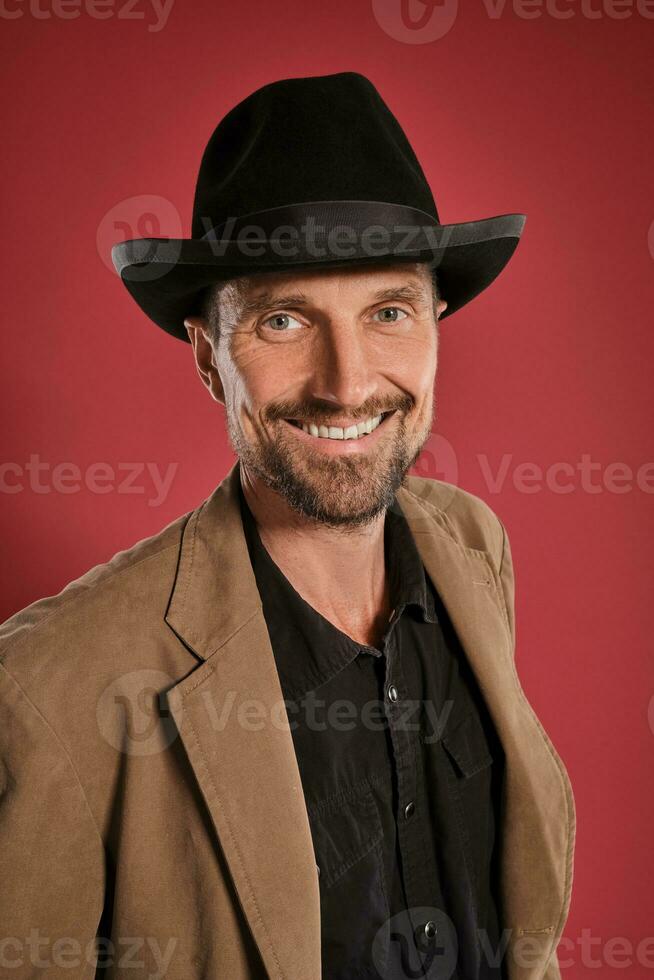 Middle-aged man with beard and mustache, wears black hat and brown jacket posing against a red background. Sincere emotions concept. photo