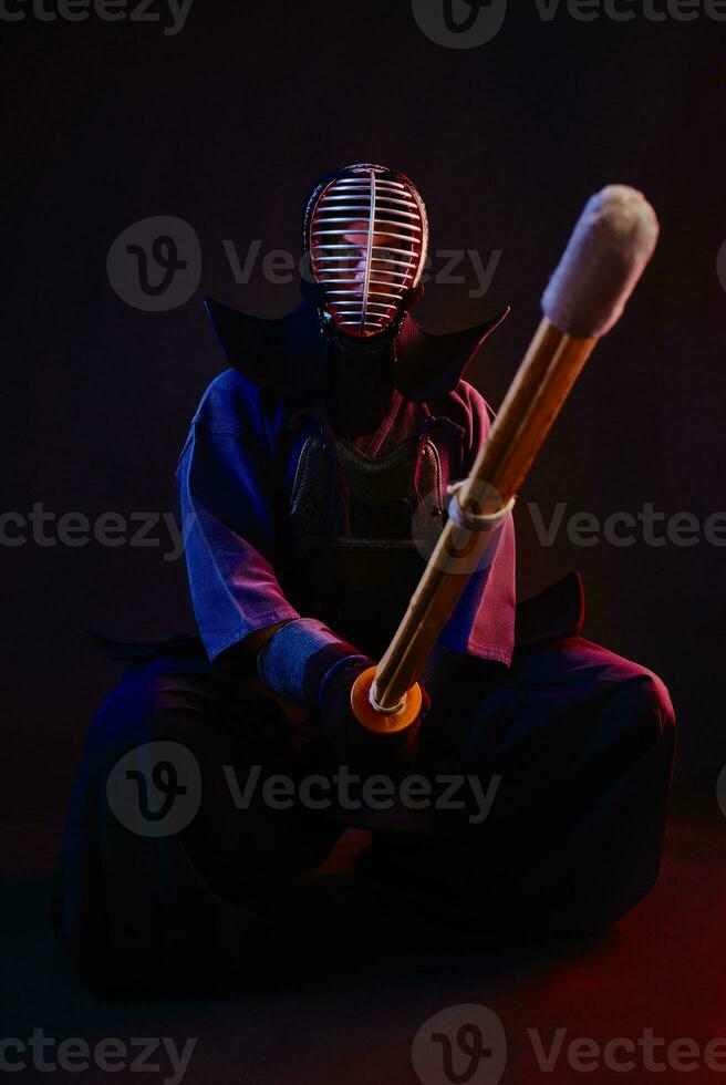 Close up. Kendo fighter wearing in an armor, traditional kimono, helmet, sitting, practicing martial art with shinai bamboo sword, black background. photo