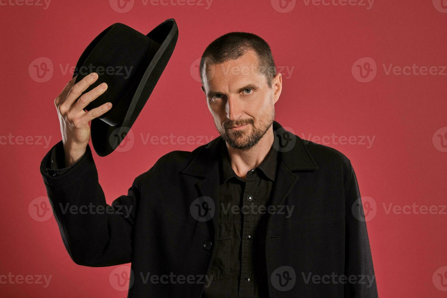 Middle-aged man with beard and mustache, wears black hat and jacket posing against a red background. Sincere emotions concept. photo