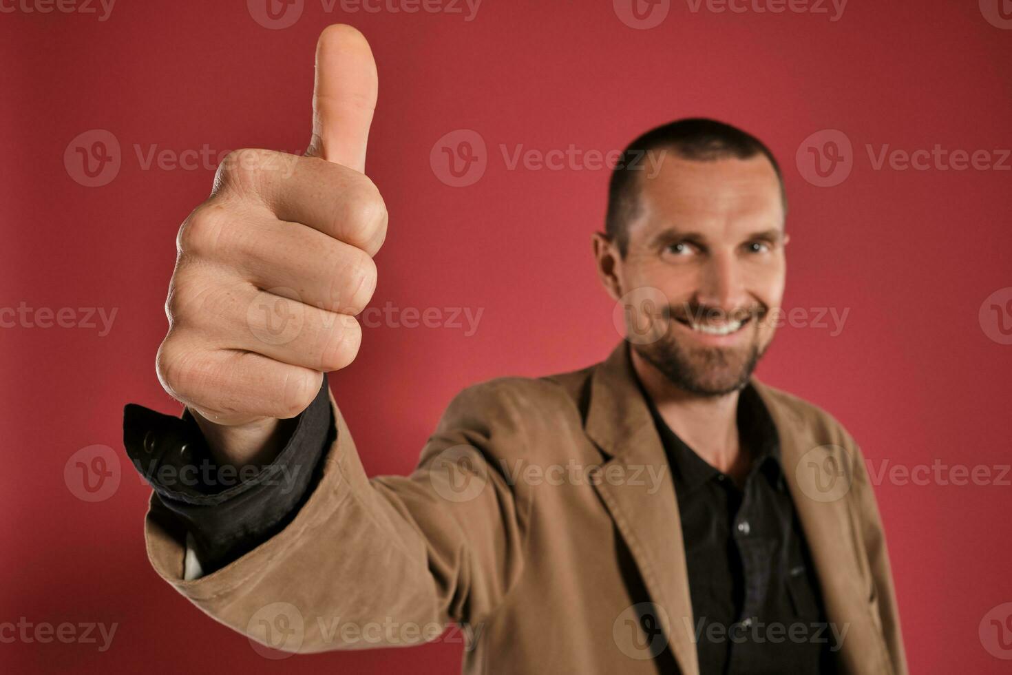 Middle-aged man with beard and mustache, wears black shirt and brown jacket posing against a red background. Sincere emotions concept. photo