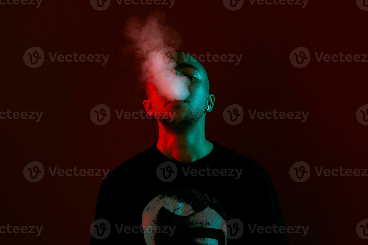 Close-up studio shot of a young bald guy vaping, blowing out a cloud of smoke on red background. photo