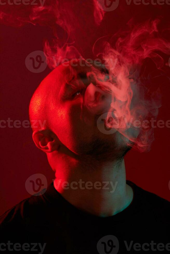 Close-up studio shot of a young bald guy vaping, blowing out a cloud of smoke on red background. photo