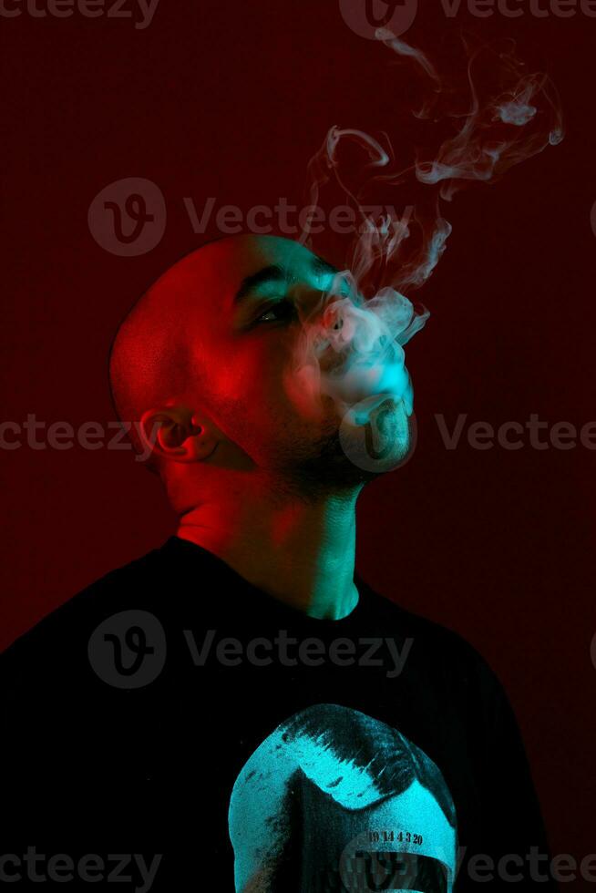 Close-up studio shot of a young bald guy vaping, blowing out a cloud of smoke on red background. photo