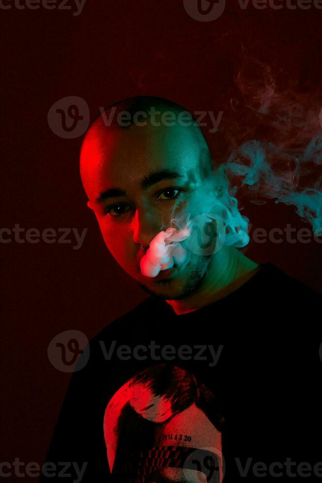 Close-up studio shot of a young bald guy vaping, blowing out a cloud of smoke on red background. photo