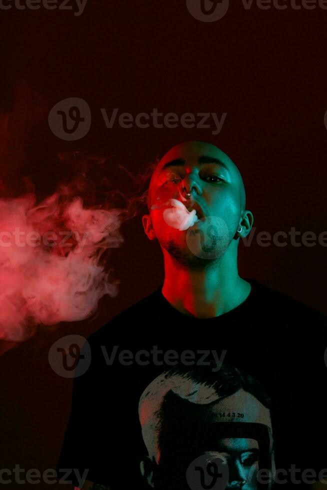 Close-up studio shot of a young bald guy vaping, blowing out a cloud of smoke on red background. photo