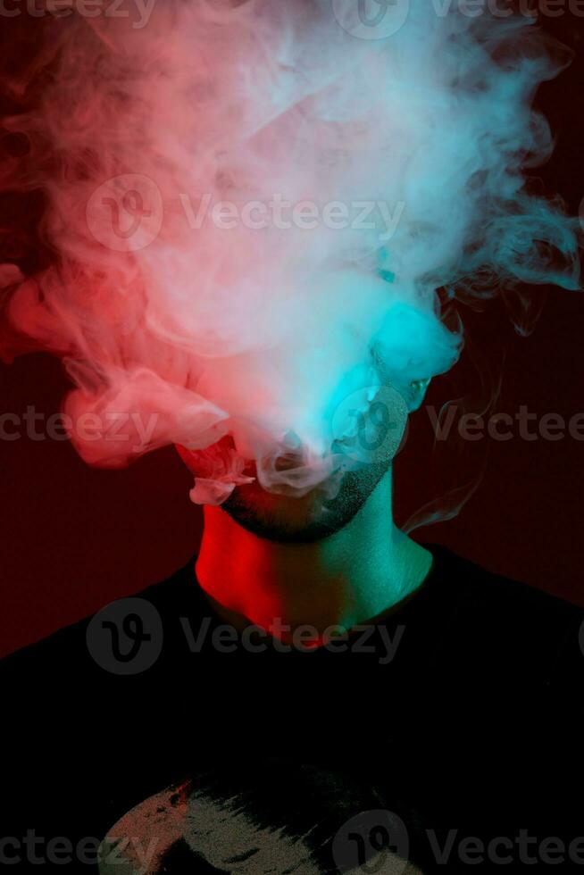Close-up studio shot of a young bald guy vaping, blowing out a cloud of smoke on red background. photo
