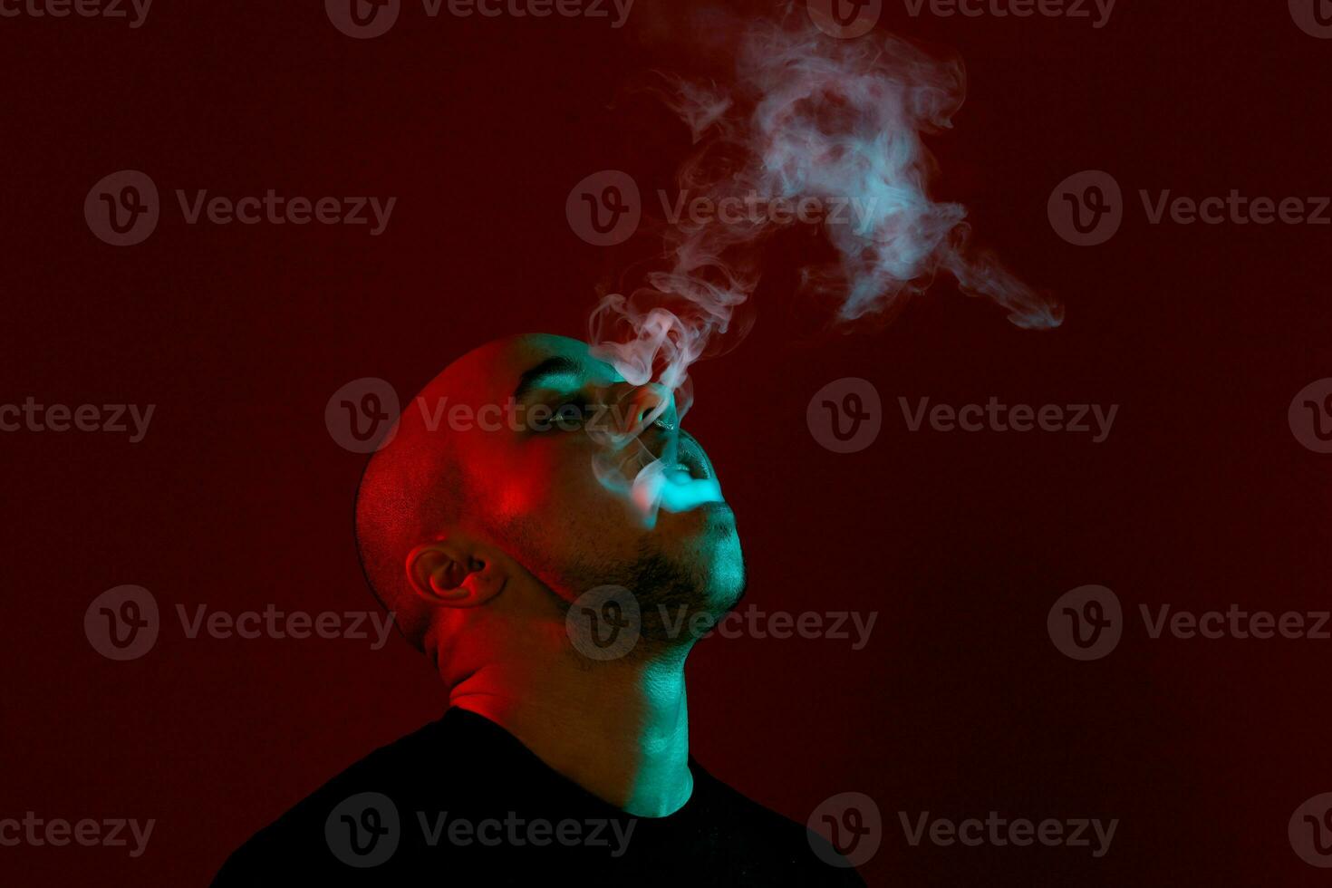 Close-up studio shot of a young bald guy vaping, blowing out a cloud of smoke on red background. photo