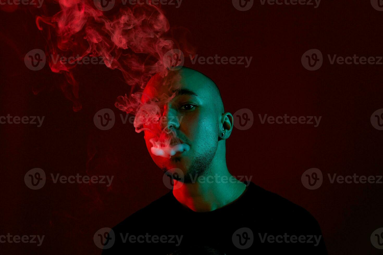 Close-up studio shot of a young bald guy vaping, blowing out a cloud of smoke on red background. photo