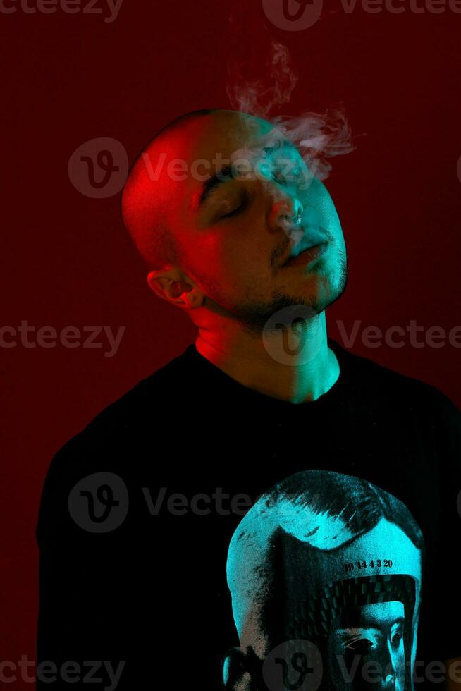 Close-up studio shot of a young bald guy vaping, blowing out a cloud of smoke on red background. photo