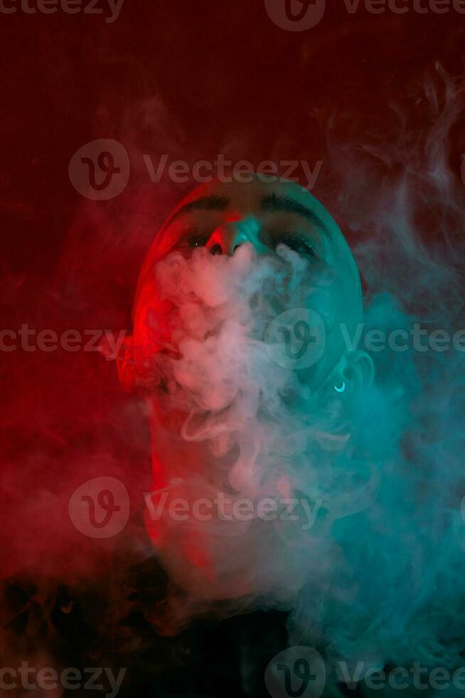 Close-up studio shot of a young bald guy vaping, blowing out a cloud of smoke on red background. photo