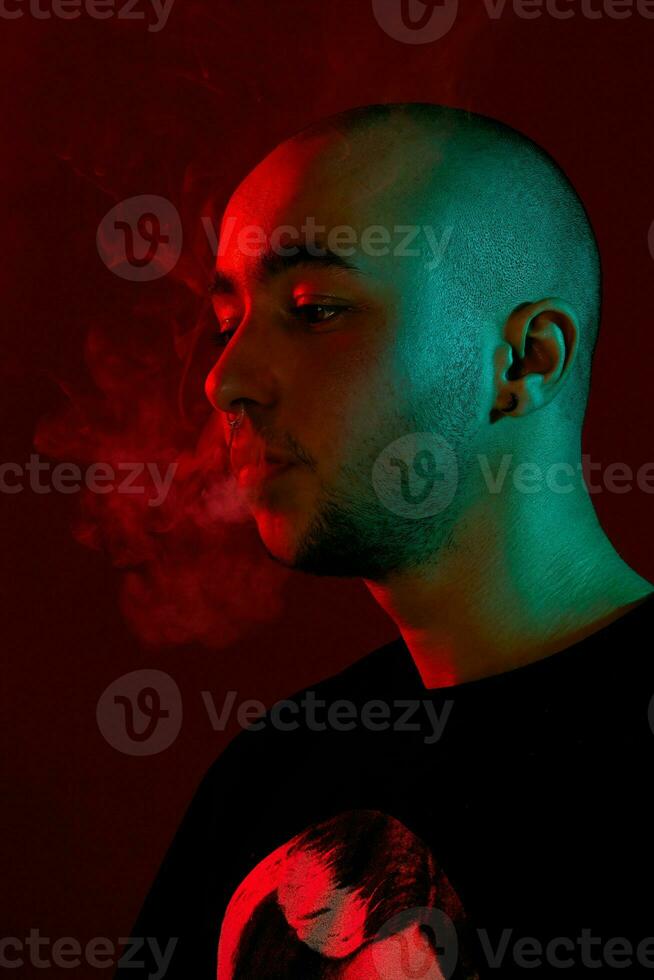 Close-up studio shot of a young bald guy vaping, blowing out a cloud of smoke on red background. photo
