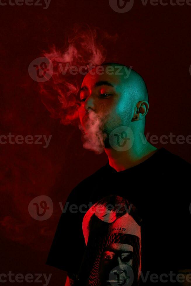 Close-up studio shot of a young bald guy vaping, blowing out a cloud of smoke on red background. photo