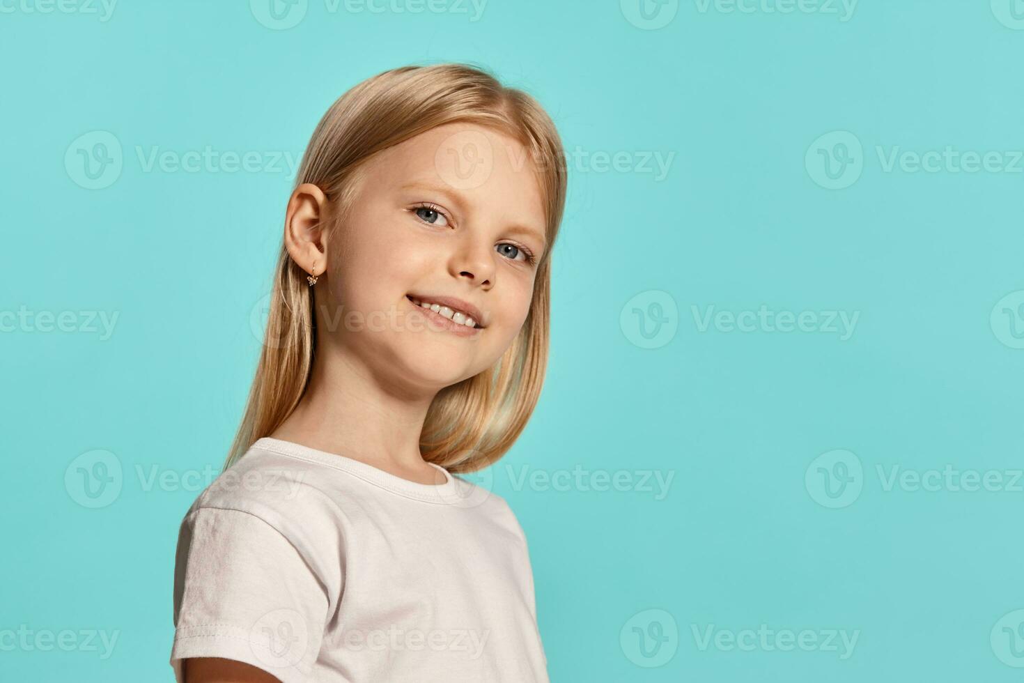 de cerca estudio Disparo de un encantador rubia pequeño niña en un blanco camiseta posando en contra un azul antecedentes. foto