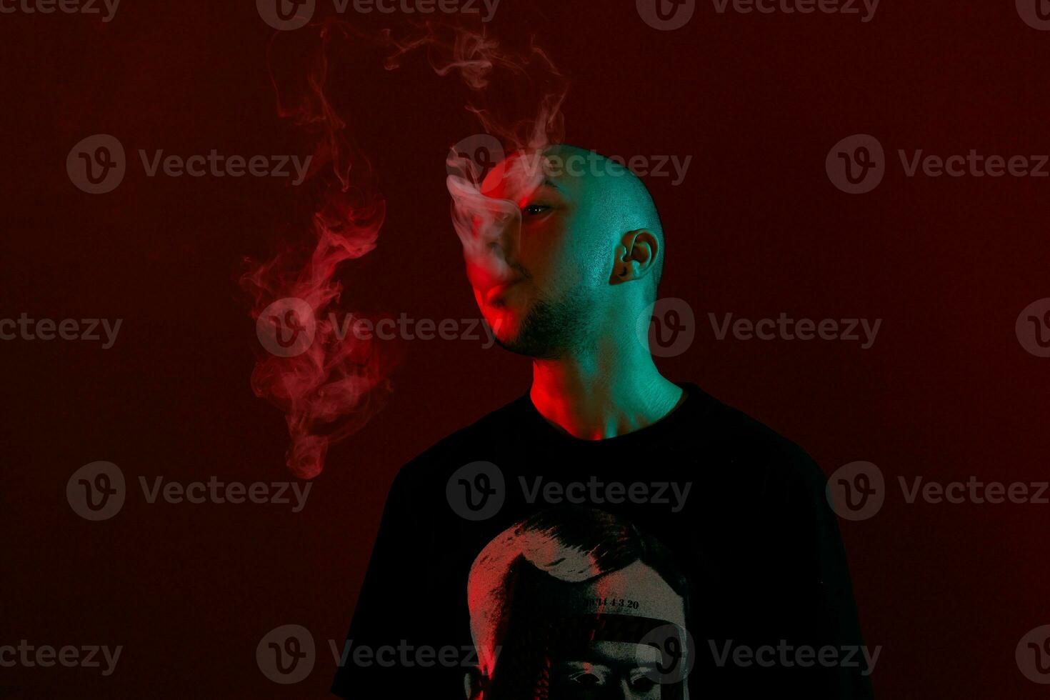 Close-up studio shot of a young bald guy vaping, blowing out a cloud of smoke on red background. photo