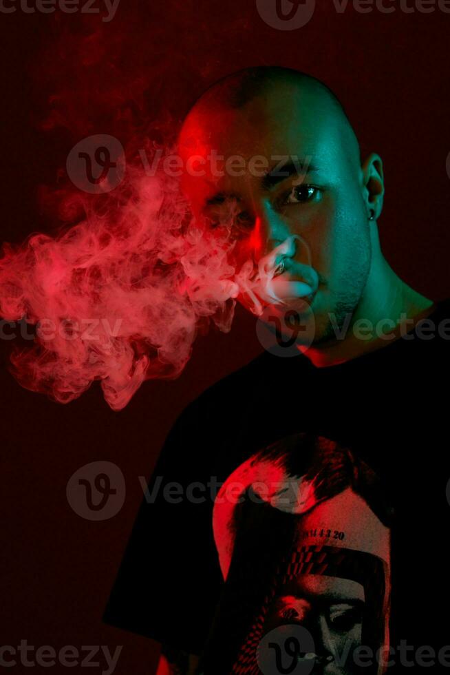 Close-up studio shot of a young bald guy vaping, blowing out a cloud of smoke on red background. photo