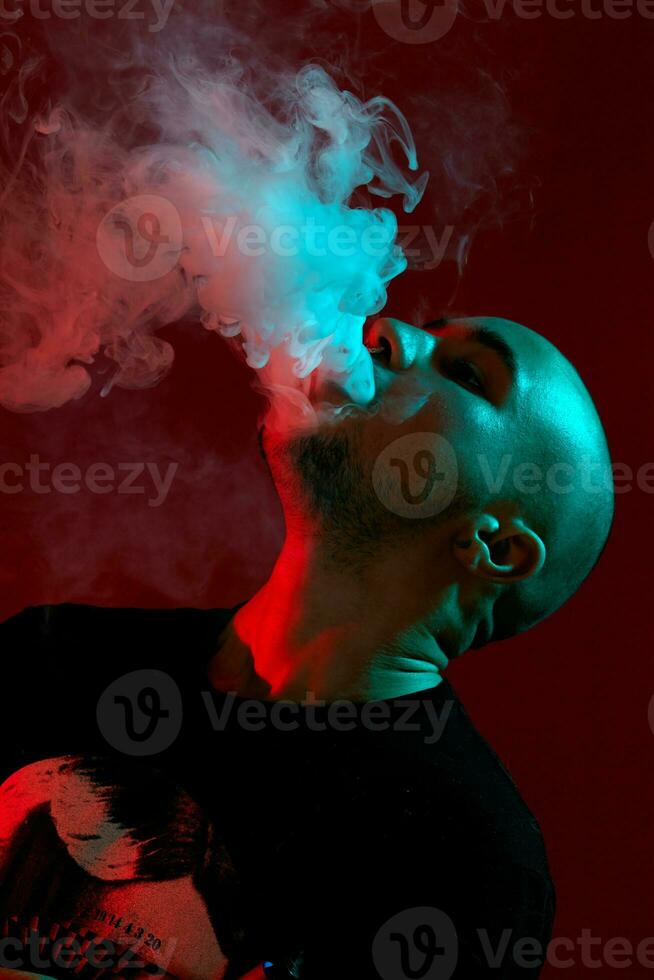 Close-up studio shot of a young bald guy vaping, blowing out a cloud of smoke on red background. photo