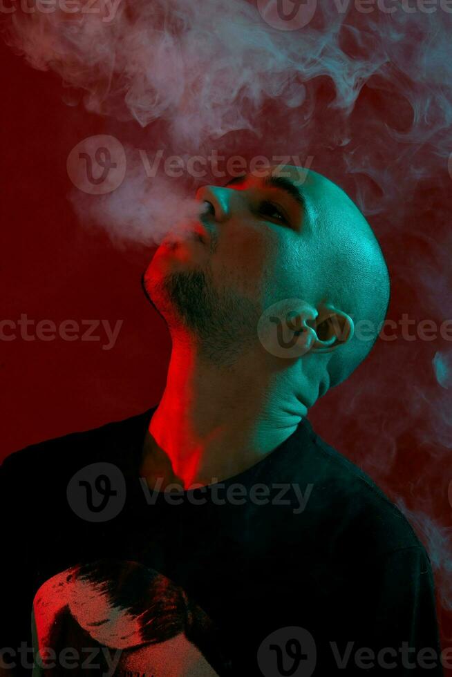 Close-up studio shot of a young bald guy vaping, blowing out a cloud of smoke on red background. photo