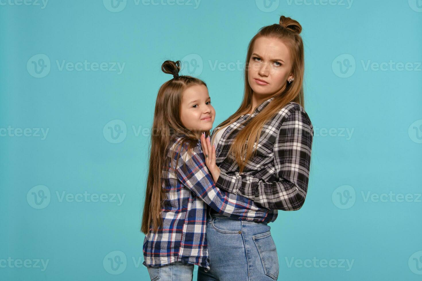 mamá y hija con un gracioso peinados, vestido a cuadros camisas y azul mezclilla pantalones son posando en contra un azul estudio antecedentes. de cerca disparo. foto