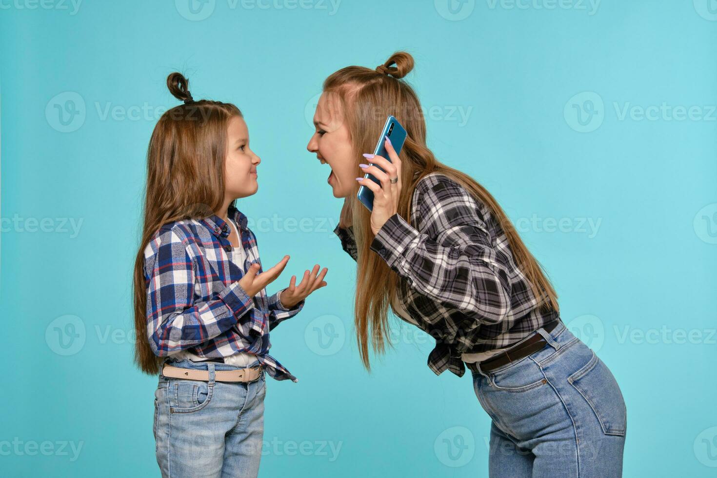 Mom and daughter dressed in checkered shirts and blue denim jeans are using smartphone while posing against a blue studio background. Close-up shot. photo