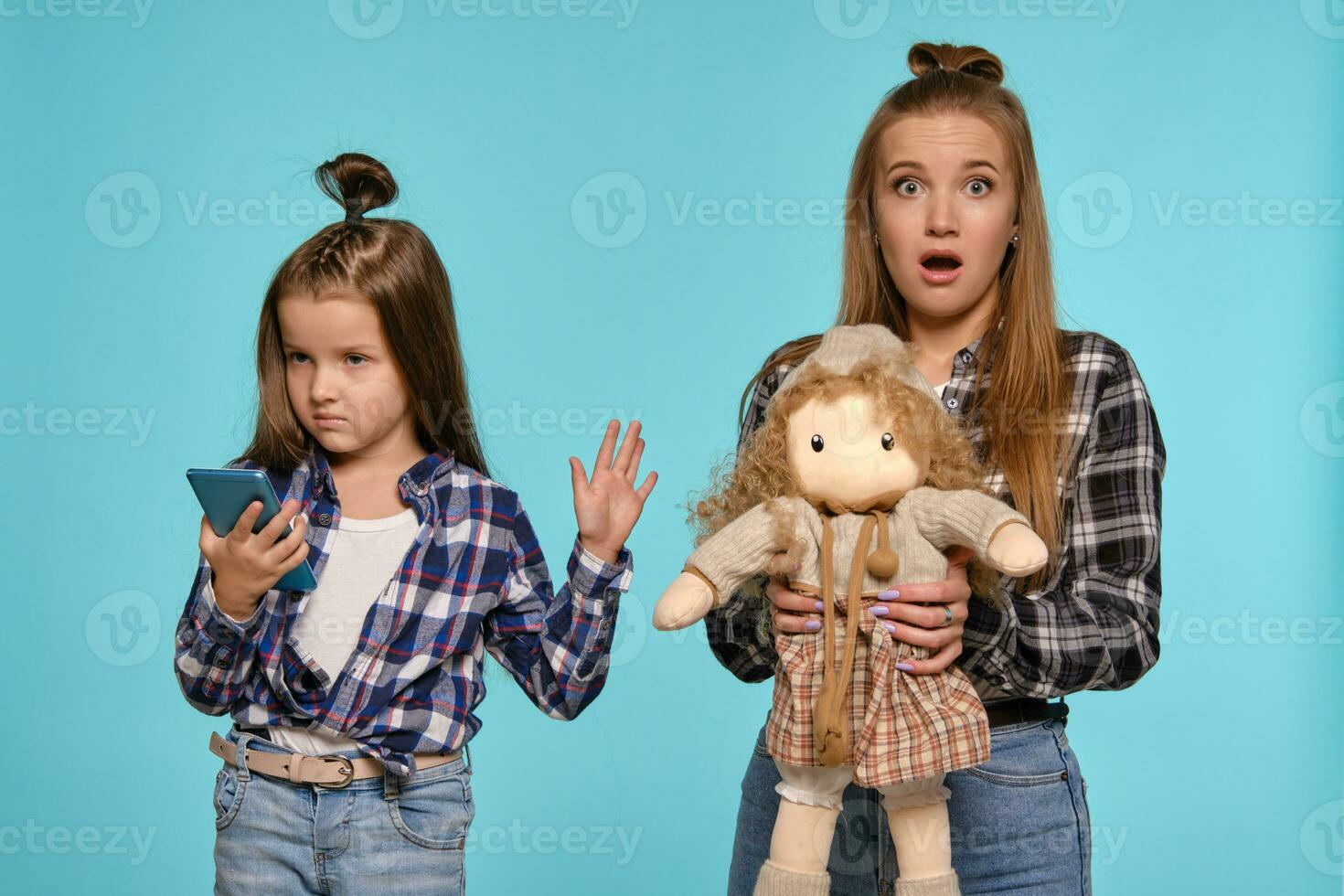 mamá y hija vestido en a cuadros camisas y azul mezclilla pantalones son utilizando teléfono inteligente mientras posando en contra un azul estudio antecedentes. de cerca disparo. foto