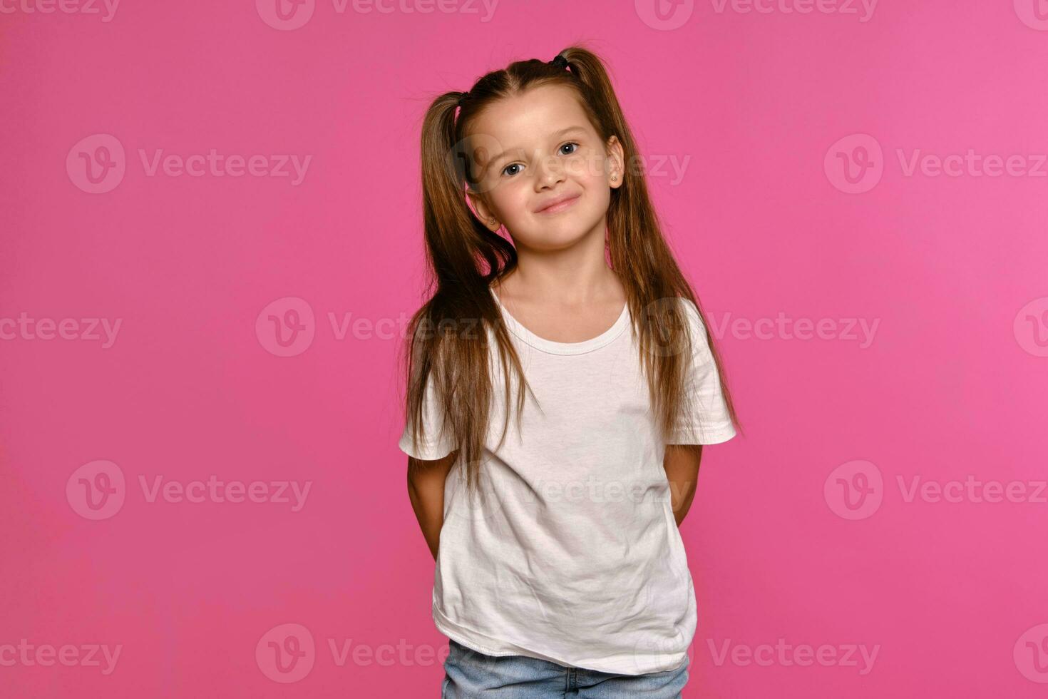 pequeño niña con colas de caballo, vestido en blanco camiseta y azul pantalones es posando en contra un rosado estudio antecedentes. de cerca disparo. sincero emociones foto