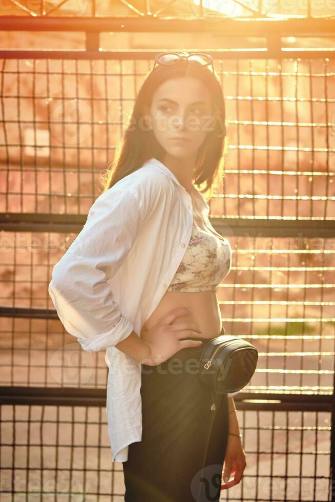 Portrait of a girl in dark sunglasses posing in city against a trellised fence. Dressed in top with floral print, white shirt, black trousers, waist bag. photo