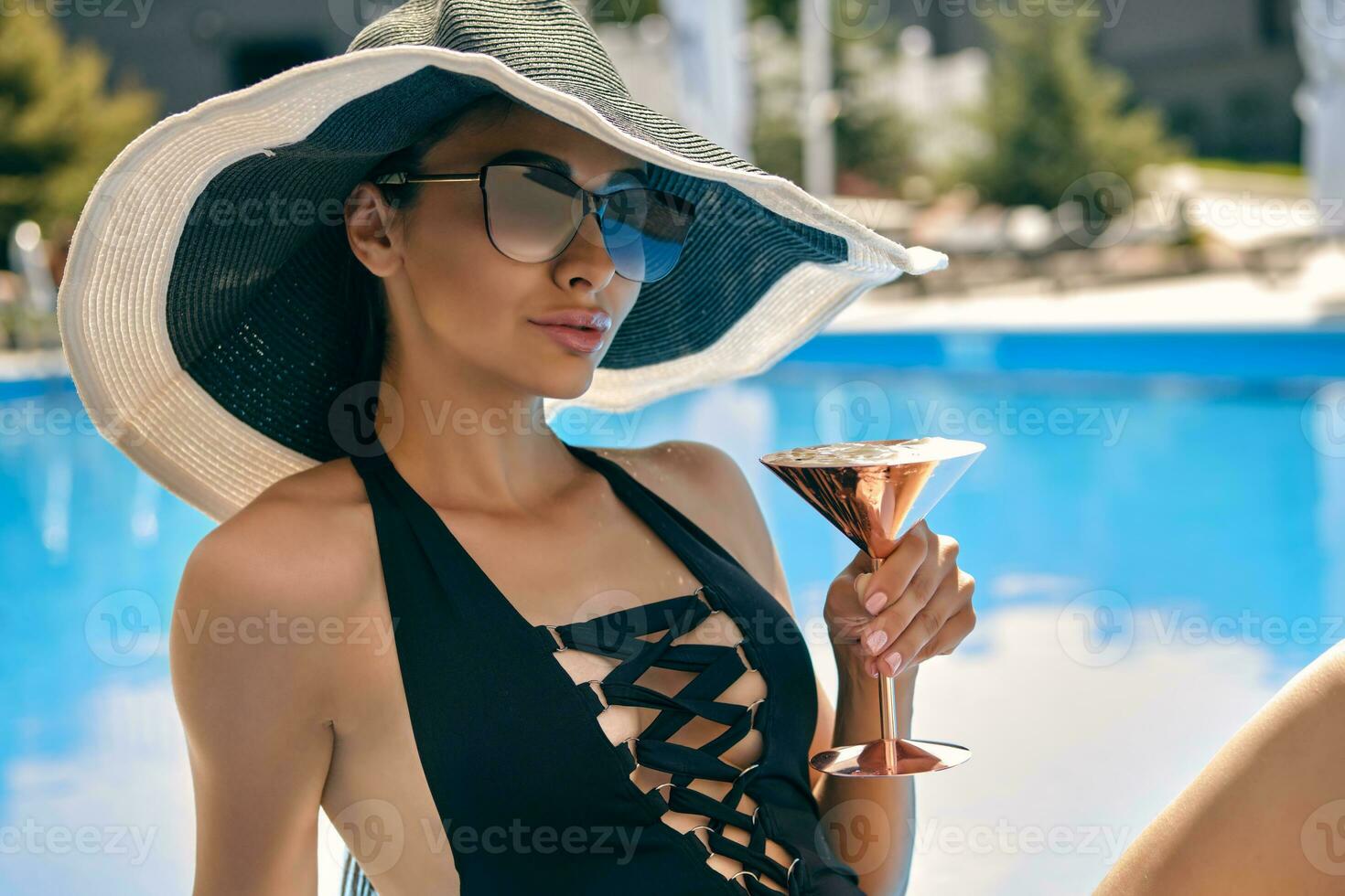 Portrait of a female having rest and posing sitting near a swimming pool. Dressed in a black swimsuit, hat and sunglasses. photo